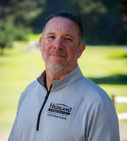 Middle-aged man with short hair and a trimmed beard wearing a light gray "Highland Commercial Roofing" pullover, standing outdoors with greenery in the background.