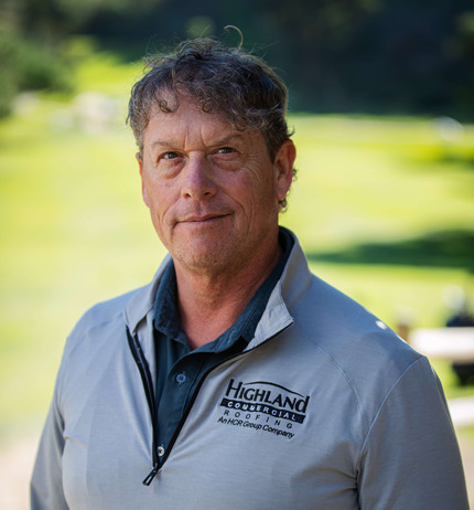 A man with curly hair wearing a light gray "Highland Commercial Roofing" jacket stands outdoors, with a blurred green background.