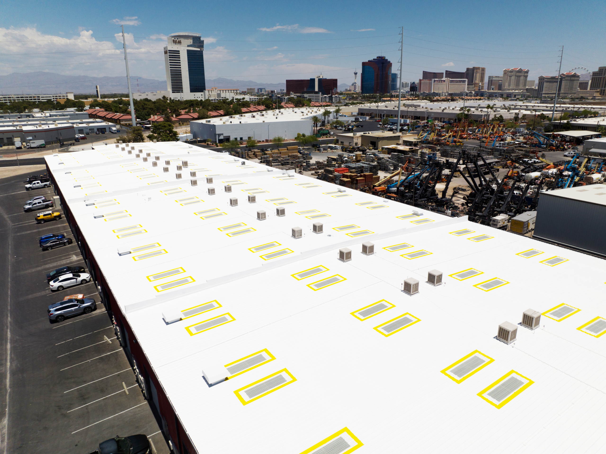 Aerial view of a large white warehouse roof with skylights and air conditioning units. Several buildings, parking lots, and a cityscape with tall buildings are visible in the background.