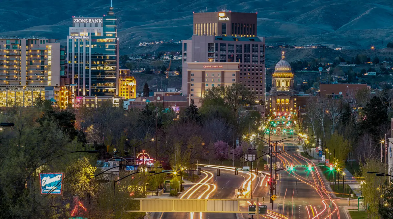 Cityscape of Boise, Idaho at dusk featuring illuminated streets and buildings including Zions Bank and the Idaho State Capitol in the background against a backdrop of hills.