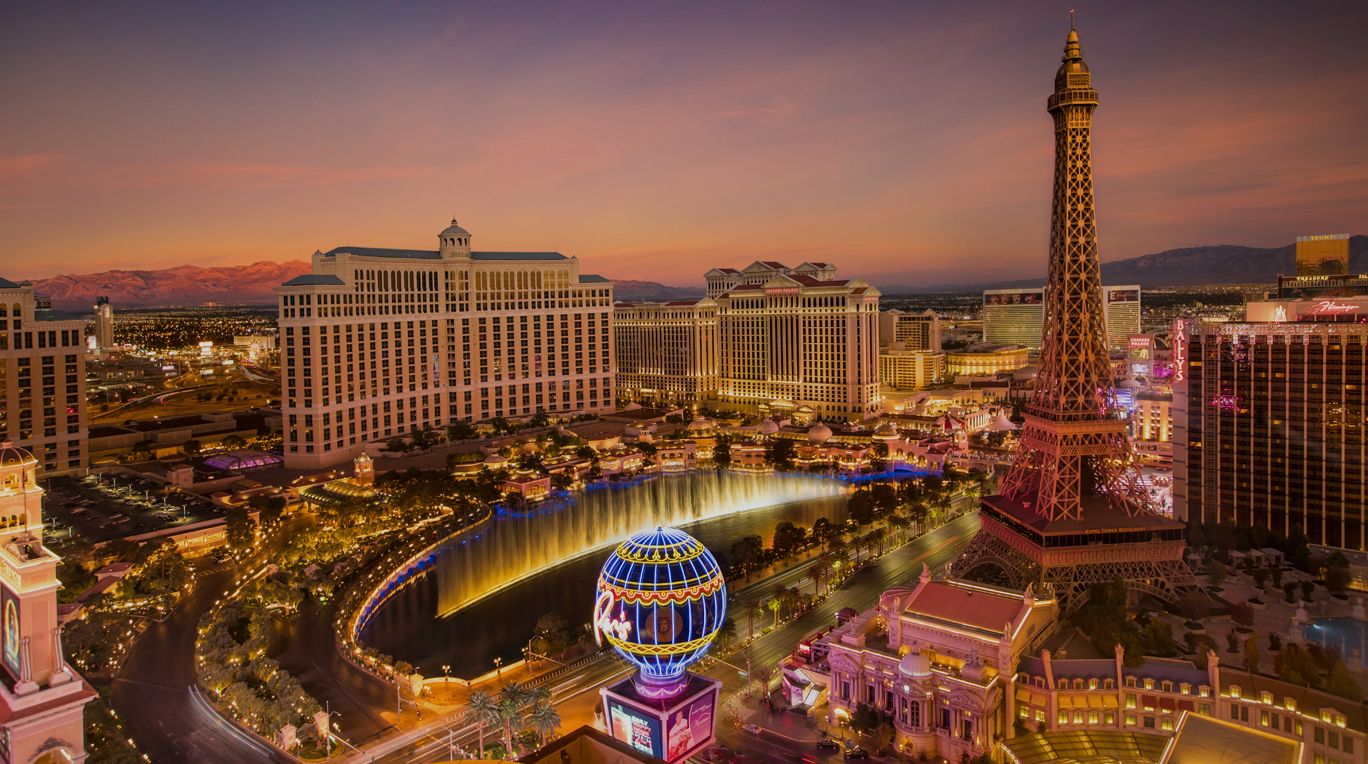 Aerial view of Las Vegas at dusk, highlighting The Bellagio with its fountain, the Paris Las Vegas Eiffel Tower, and surrounding hotels and casinos, against a backdrop of mountains and evening sky.
