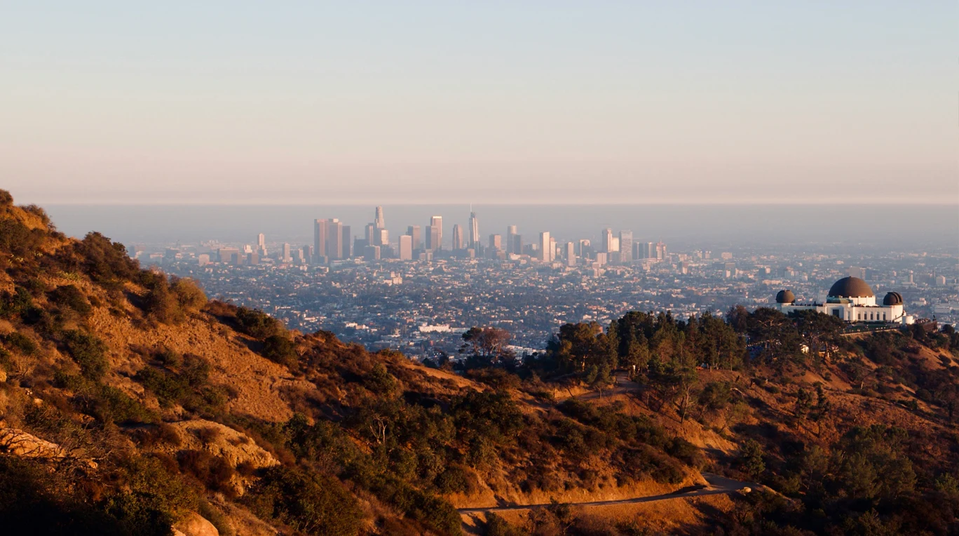 View of the Southern California cityscape from a distance, with Griffith Observatory on the right and rolling hills in the foreground. Clear sky and slight haze in the background.