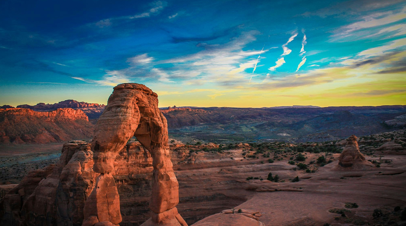 A natural stone arch stands in the middle of a rugged landscape at Arches National Park, Utah, under a vibrant sky at sunset.