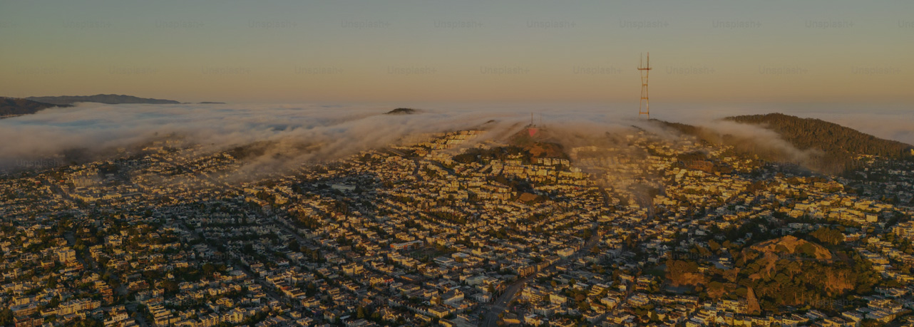 Aerial view of a sprawling cityscape with low-lying fog covering parts of the hilly terrain and scattered neighborhoods under golden hour sunlight; a tall radio tower is visible on a hill.