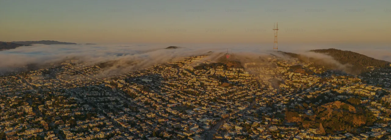 Aerial view of San Francisco's Twin Peaks at sunset, with fog rolling over the hills and a large radio tower in the background. Rows of houses and streets stretch out in the foreground, reminiscent of Southern California's sprawling landscapes.