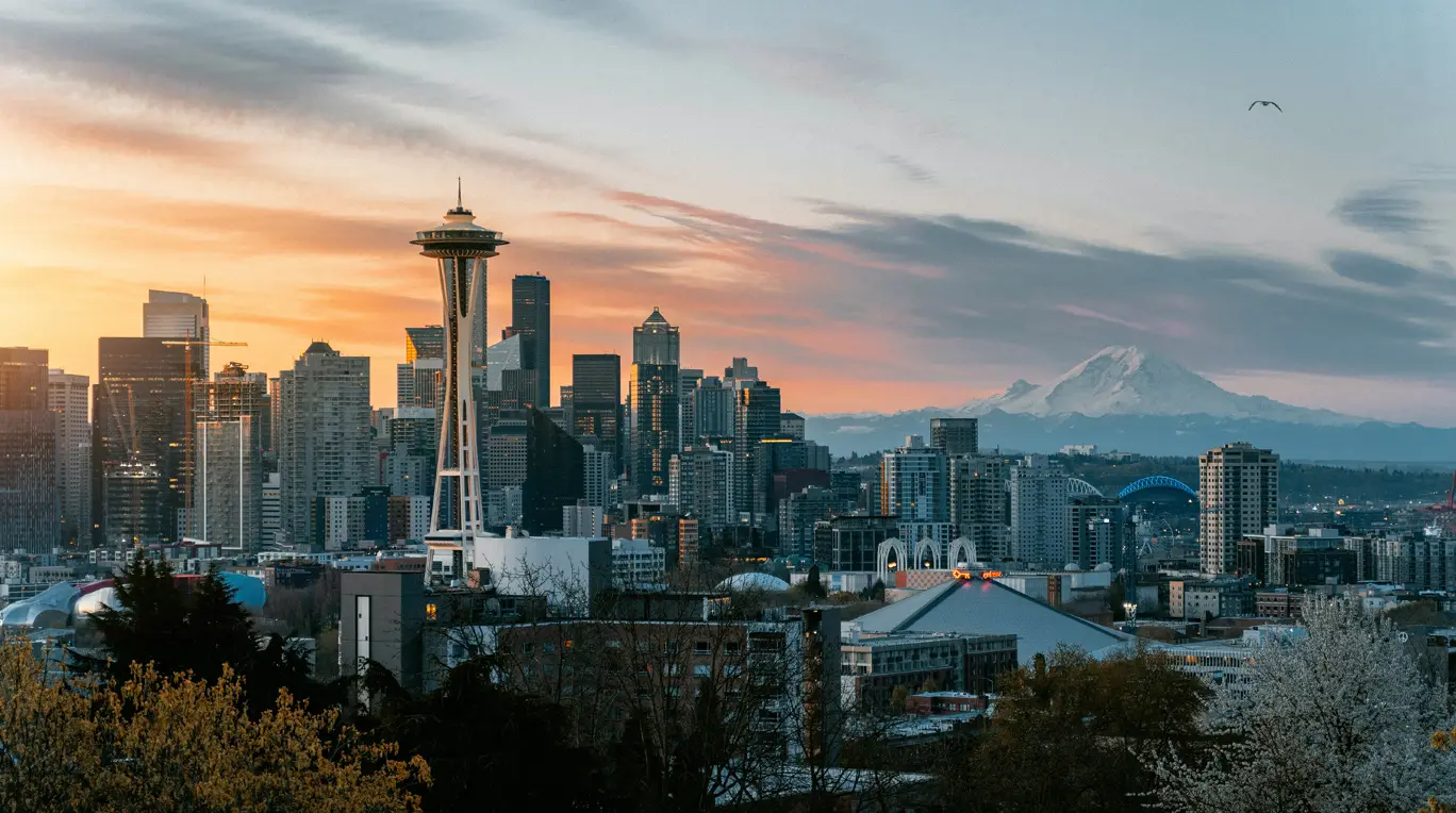 Seattle cityscape at sunset featuring Space Needle, with Mount Rainier visible in the background and a bird flying in the sky.