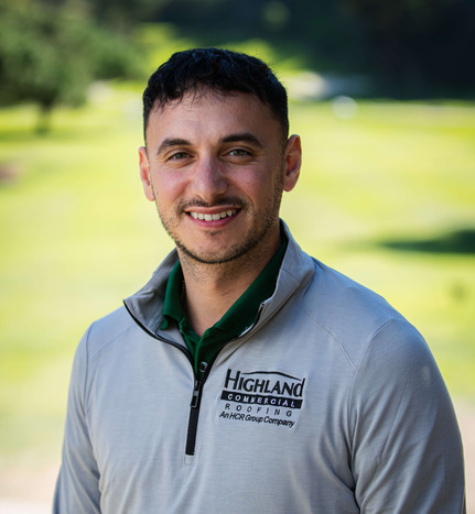 A man wearing a light gray Highland Commercial Roofing jacket stands outdoors, smiling with a blurred green landscape in the background.
