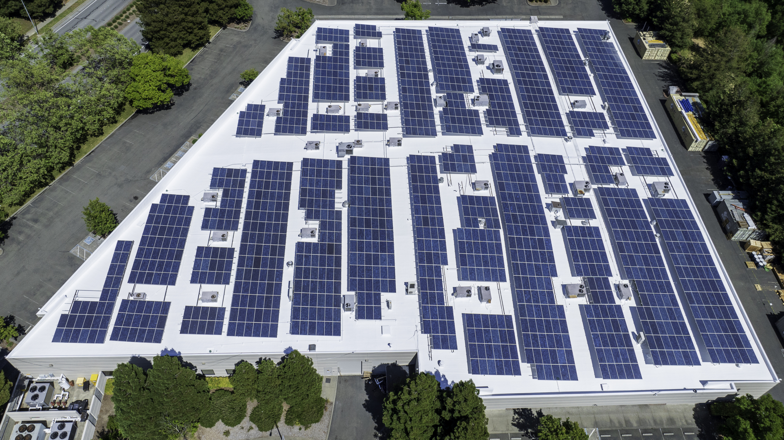 Aerial view of a large rooftop covered with solar panels surrounded by trees and parking areas.