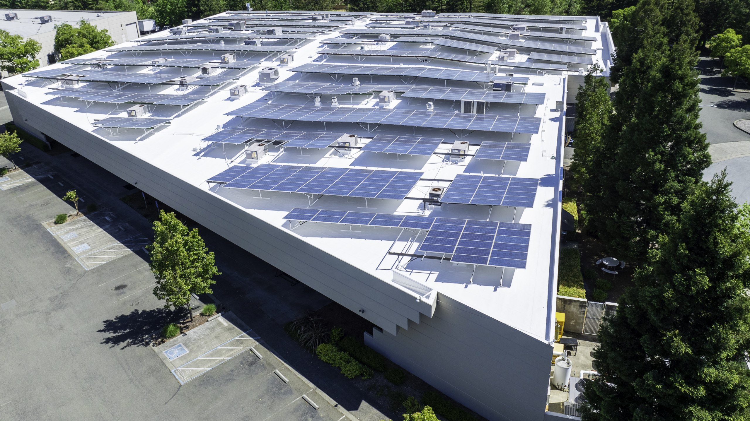 Aerial view of a large building's rooftop covered with numerous solar panels. The surrounding area includes trees and a parking lot.