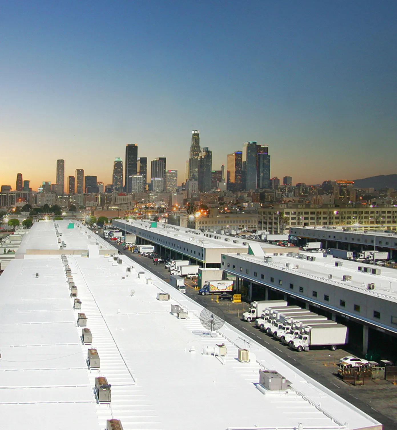 Rows of industrial warehouses with white roofs and parked trucks in the foreground, with a city skyline and clear sky in the background.