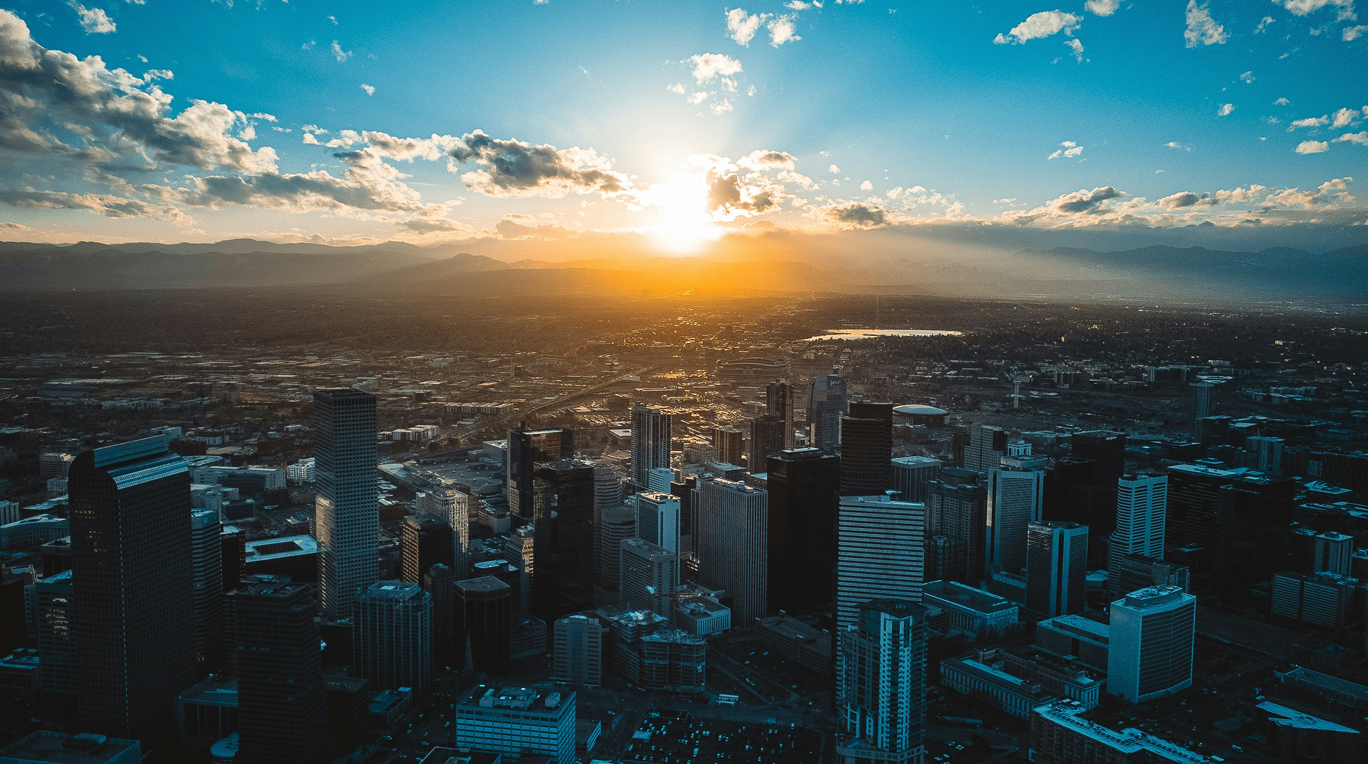 Aerial view of a city skyline at sunset with skyscrapers in the foreground and mountains in the distance under a partly cloudy sky.