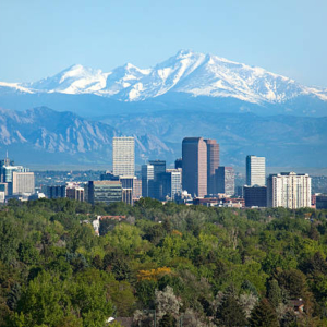 City skyline with tall buildings surrounded by greenery, against a backdrop of snow-capped mountains under a clear blue sky.