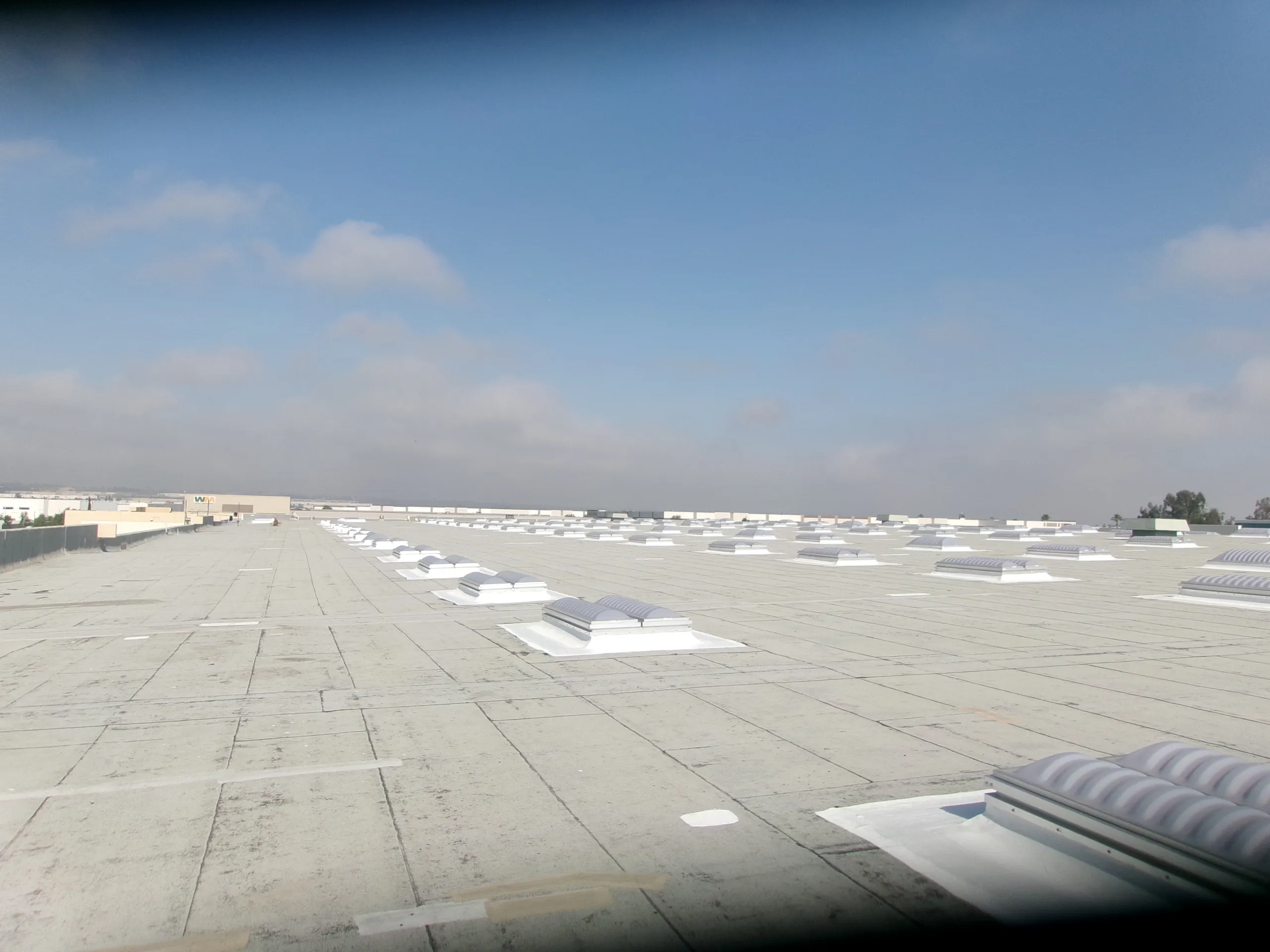Flat industrial roof with multiple rectangular skylights under a mostly clear blue sky with some clouds, viewed from one corner at an angle.