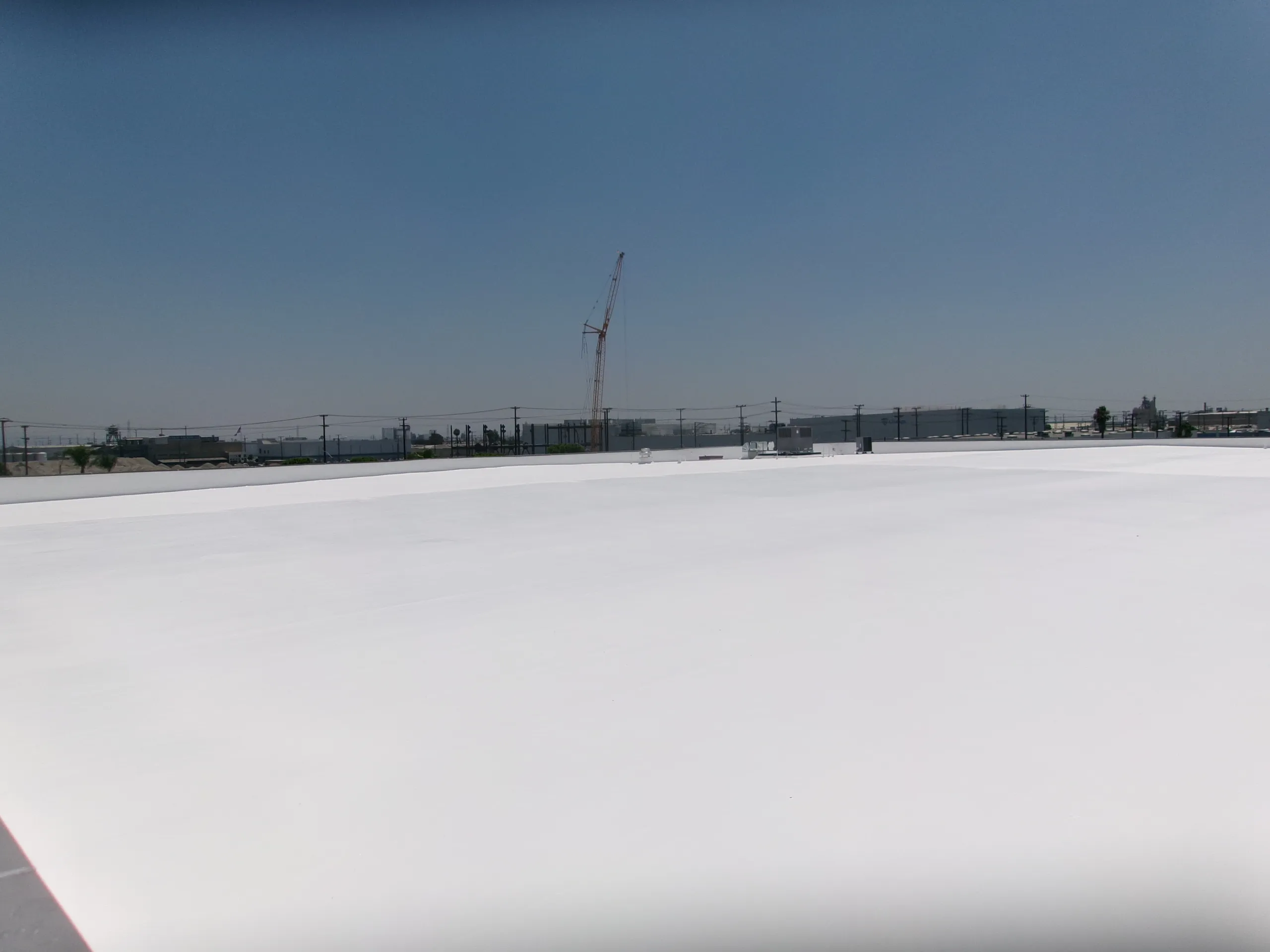 A large, flat white roof under a clear sky with a crane and industrial buildings visible in the distant background.