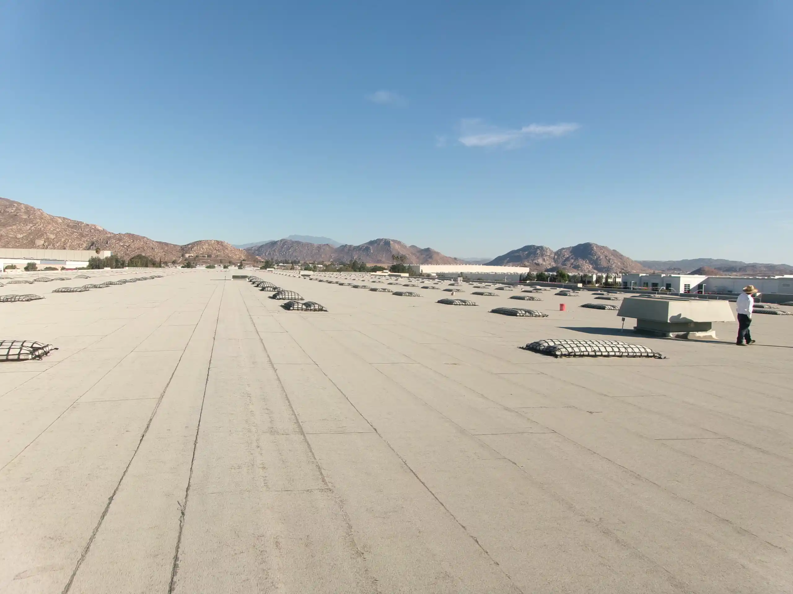 Flat industrial rooftop with rows of large vent covers and a person standing on the right, showcasing one of the top flat roofing systems for commercial buildings, with mountains and a clear blue sky in the background.
