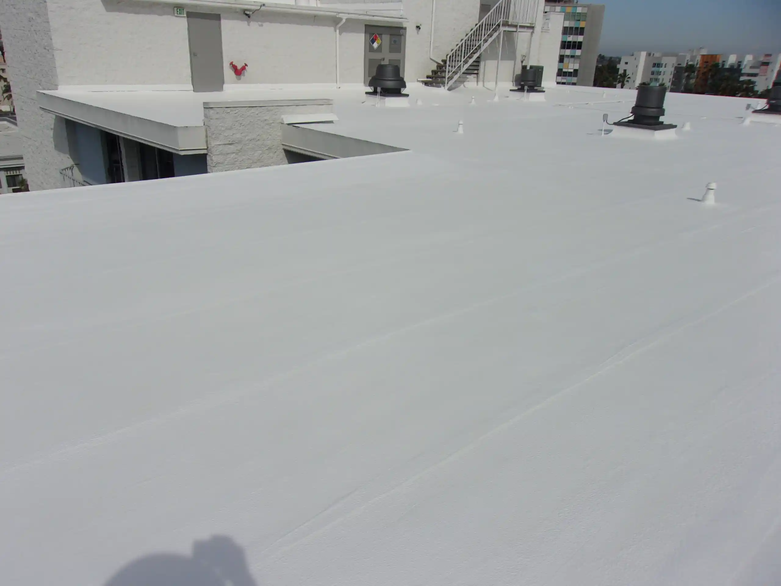 Flat white rooftop with several ventilation units, white-painted buildings, and external metal stairs in the background under a clear sky, demonstrating the importance of choosing the best flat roof repair material to maintain durability and a clean appearance.