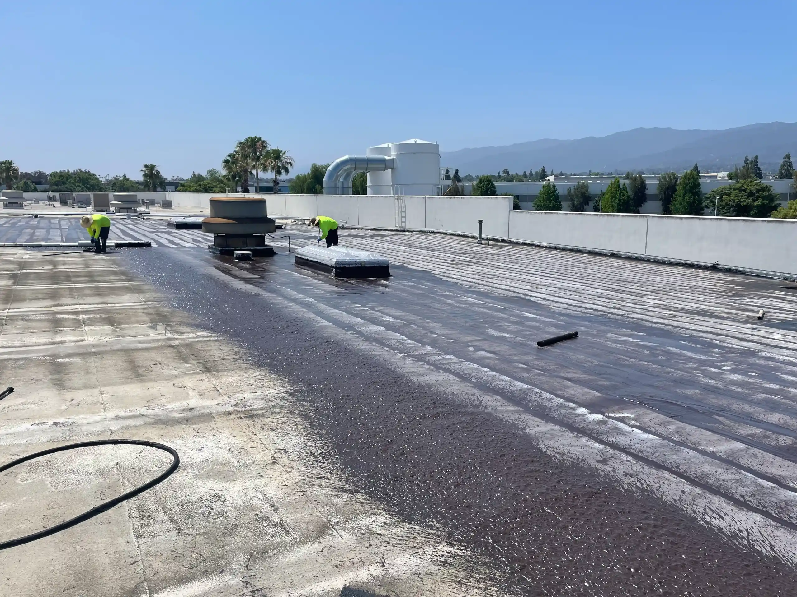 Workers are applying a dark coating to a flat commercial rooftop, one of the common types of commercial roofing systems, on a clear day, with HVAC units and mountains visible in the background.