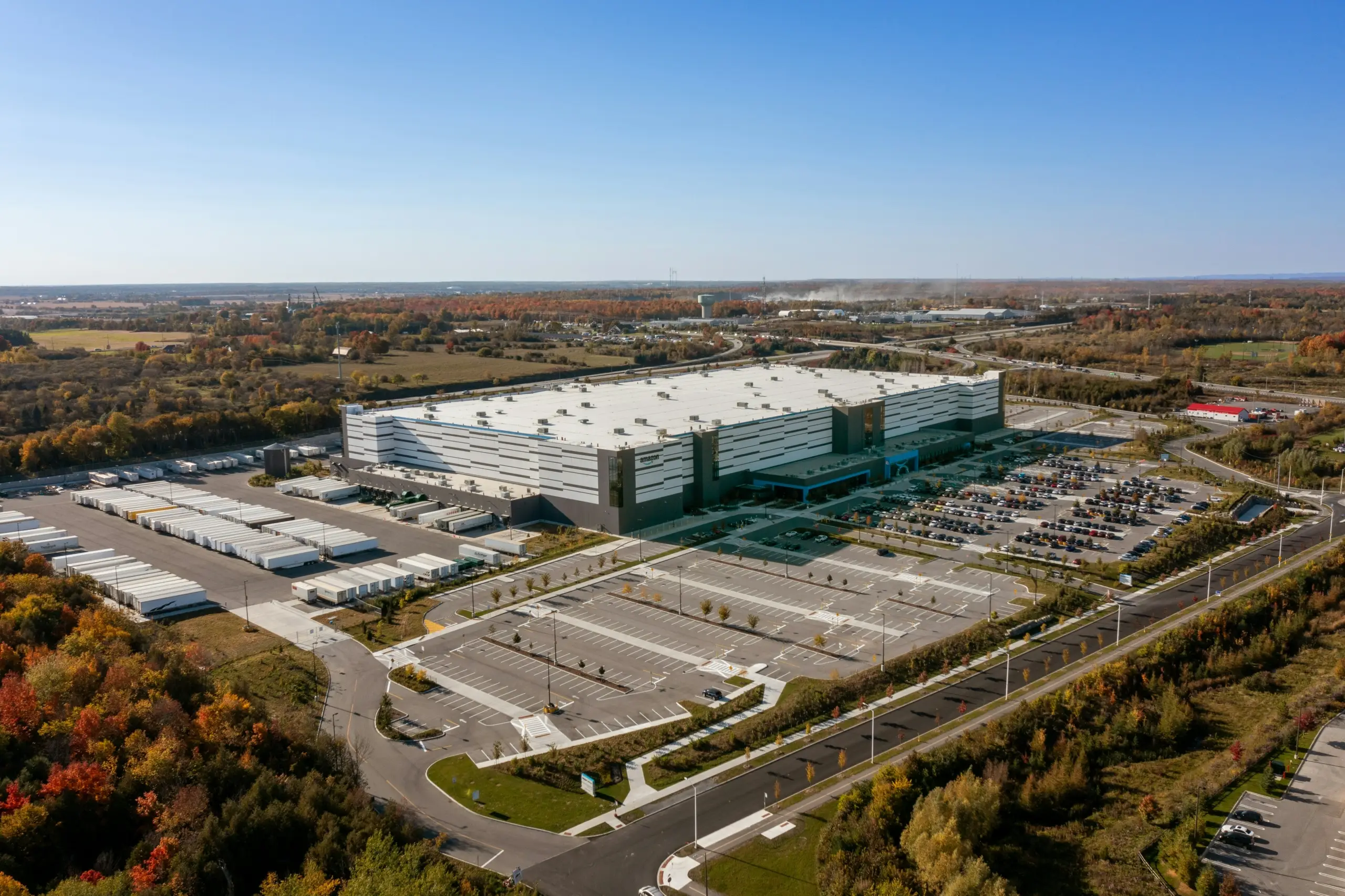 Aerial view of a large warehouse or distribution center with surrounding parking lots, loading docks, and nearby trees during daytime.