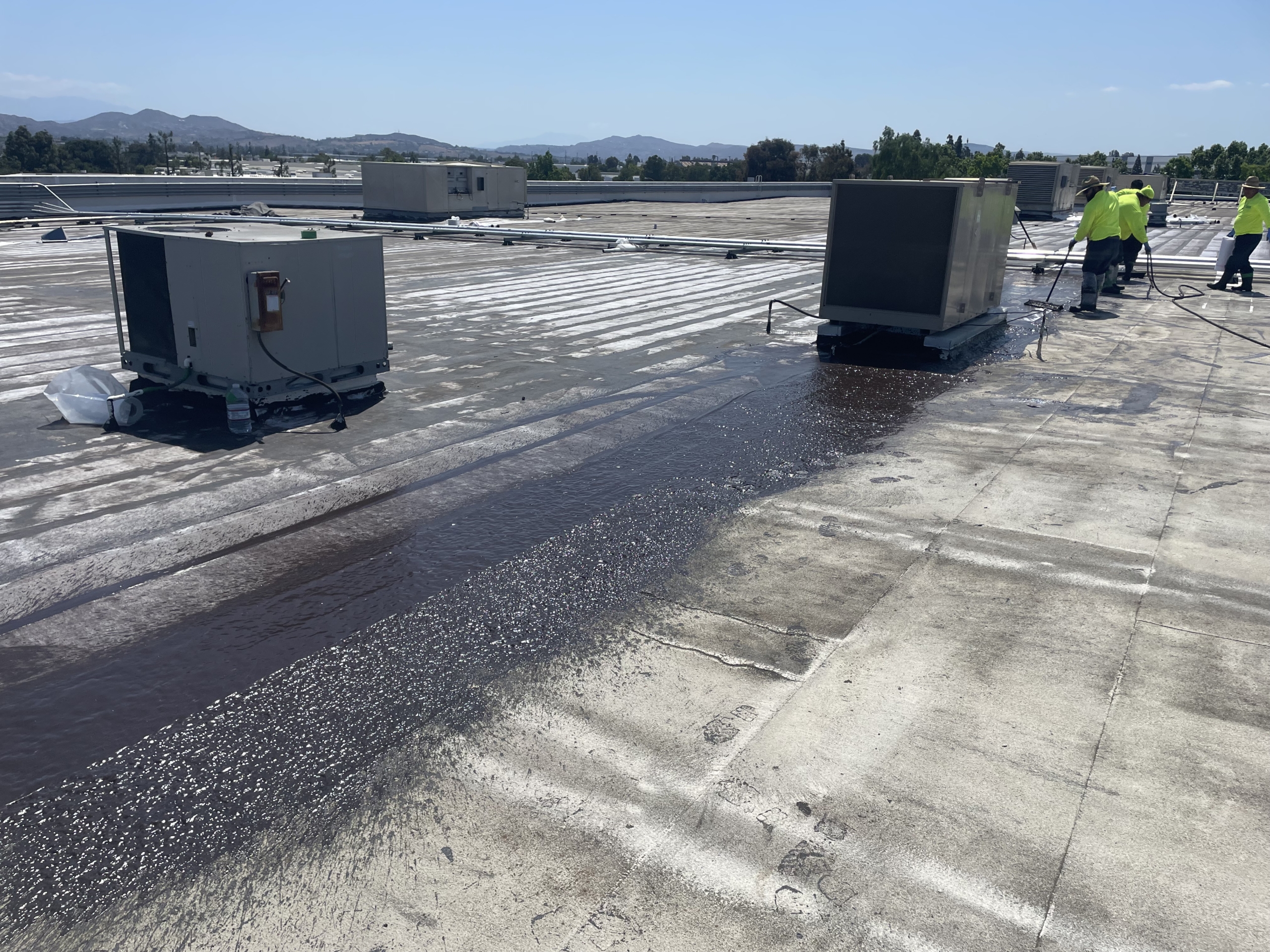 Workers in yellow vests apply protective coating to a flat commercial rooftop near HVAC units on a sunny day, mindful of what are the effects of El Niño and how changing weather patterns can impact building maintenance.