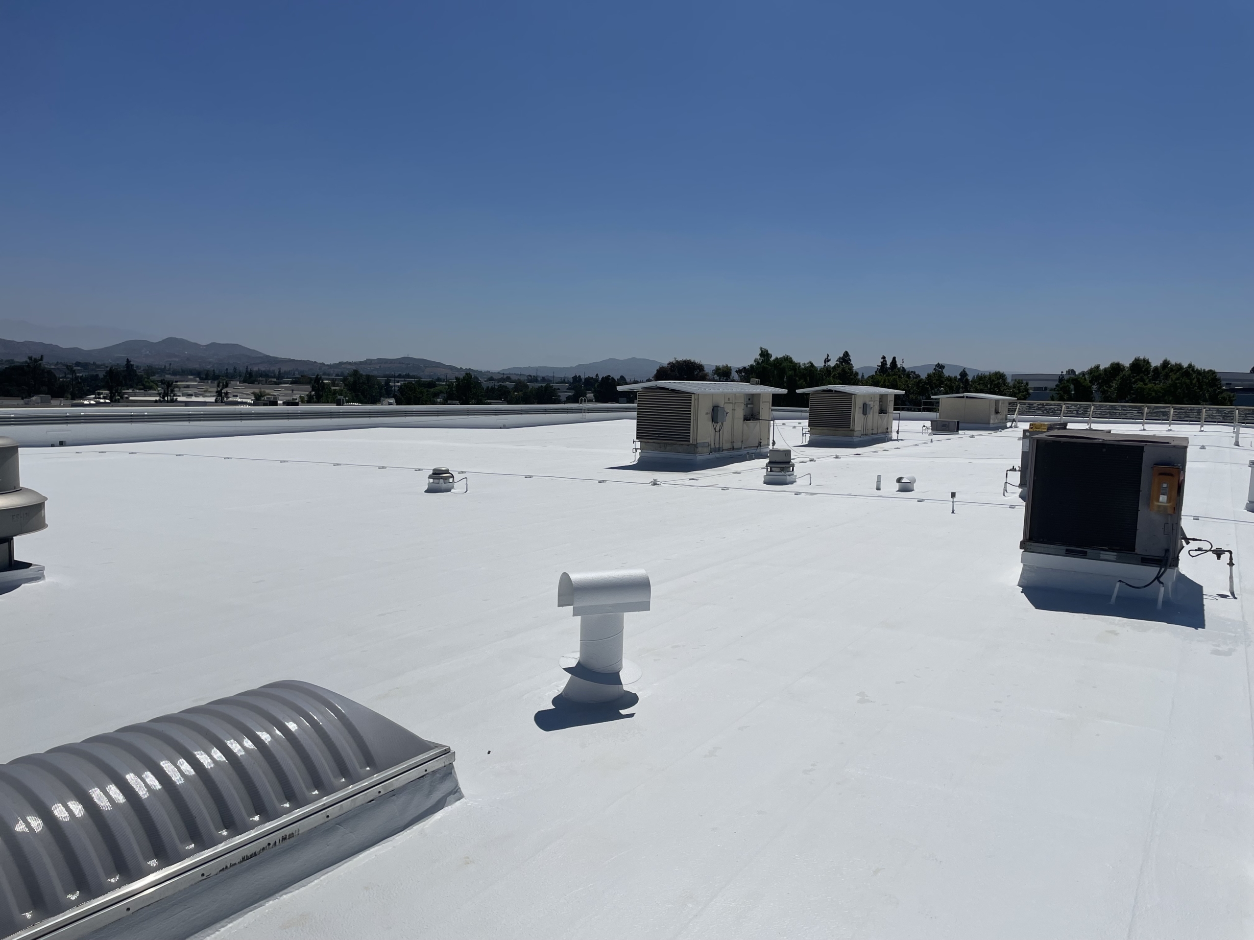 Flat white rooftop with multiple HVAC units, vents, and skylights under a clear blue sky; distant hills and trees are visible in the background. Featuring TPO roofing, this building showcases durability and efficiency—an important factor for property owners wondering how long does a TPO roof last.